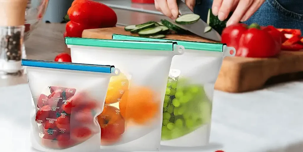 Various reusable silicone food storage bags filled with tomatoes, oranges, and grapes placed on a kitchen counter while vegetables are being cut in the background.