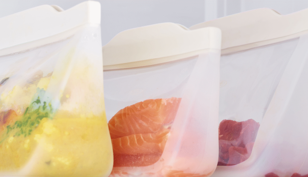 Reusable silicone food storage bags containing soup, salmon, and meat pieces.